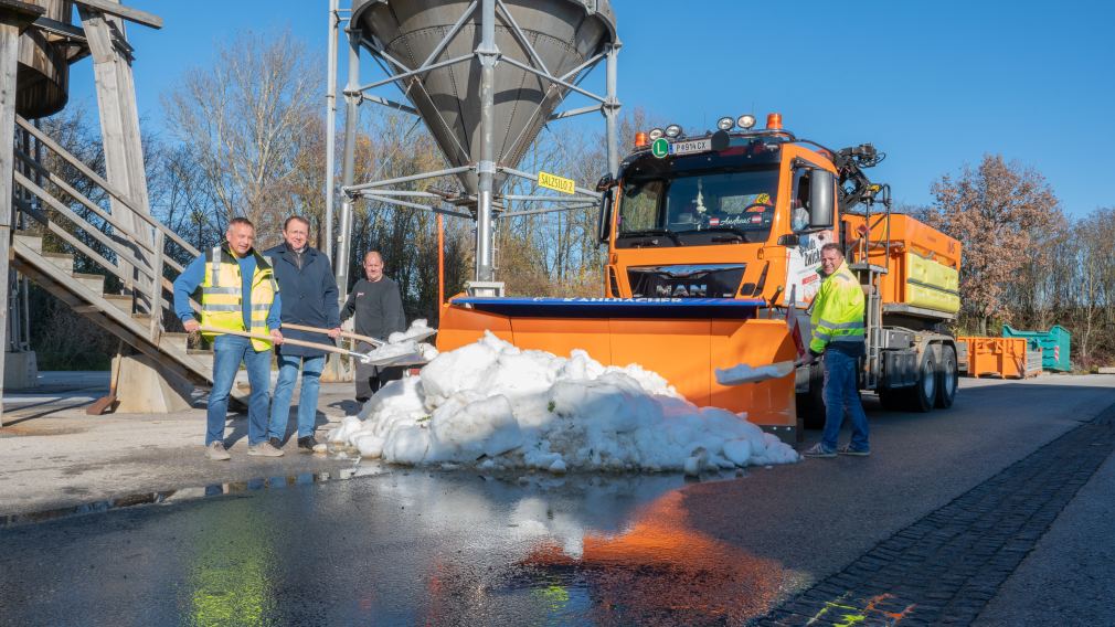 Mehrere Männer stehen mit einer Schneeschaufel in der Hand bei einem Haufen Schnee, im Hintergrund befindet sich ein Schneepflug