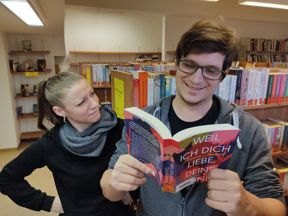 Zwei Menschen stehen in der Stadtbücherei mit einem Buch in der Hand. (Foto: Stadtbücherei)