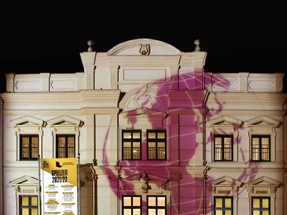 Das beleuchtete Landestheater Niederösterreich bei Nacht ist zu sehen. Auf die Fassade ist eine violette Weltkugel projiziert und eine Fahne mit der Aufschrift 