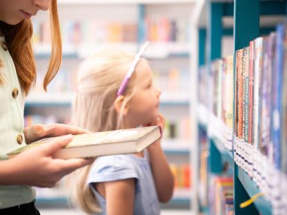 Ein Buch in der Hand eines Mädchens, verschwommen im Hintergrund schmökert ein jüngeres Mädchen durch die Bücher der Bücherei.