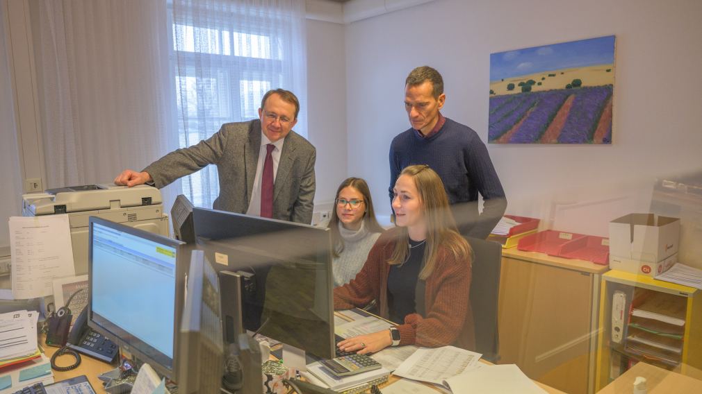 Zwei Damen und Zwei Herren stehen bzw. sitzen an einem Schreibtisch und schauen in einen Computerbildschirm.