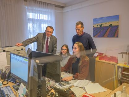 Zwei Damen und Zwei Herren stehen bzw. sitzen an einem Schreibtisch und schauen in einen Computerbildschirm.